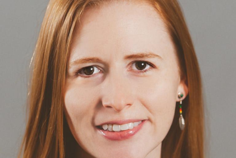 The image shows a woman with long, straight red hair wearing a green top. She has a friendly expression with a slight smile and is facing directly at the camera. She is wearing dangling earrings. The background is a plain light gray. She has light skin and an approachable demeanor.