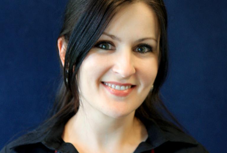 The image shows a woman with long, dark hair styled in a low ponytail with some hair framing her face. She is wearing a dark-colored top with a collar and a necklace with large, red geometric beads. She has a friendly expression and a slight smile. The background is a solid, deep blue color. She has light skin and dark eyes.