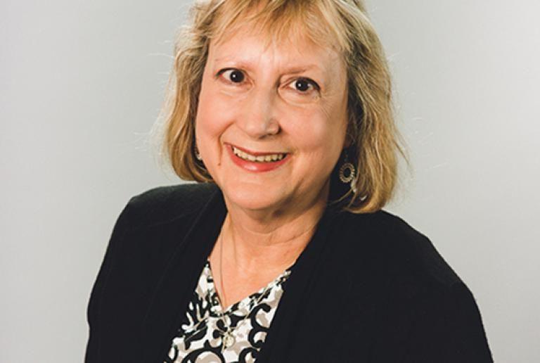 The image shows a woman with light-colored hair, styled in a shoulder-length cut with bangs. She is wearing a black cardigan over a patterned blouse with black, white, and gray colors. She has a friendly expression, and her lips are lightly colored with lipstick. The background is a plain light gray. She is also wearing hoop earrings.